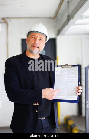 Confident architect showing building plan or report, wearing helmet and business attire on construction site. Copy space, moc up Stock Photo