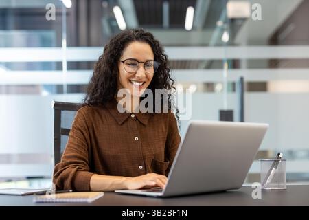 A cheerful woman with glasses works at a desk in a modern office, focused on her laptop with a big smile. The office is bright Stock Photo