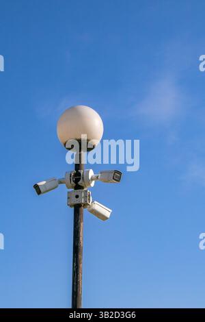 A street lamp with a round white bulb is mounted on a pole, surrounded by multiple surveillance cameras. The clear blue sky serves as a backdrop, indi Stock Photo