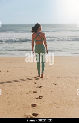 rear view of a woman walking down a beach to the sea, footprints in the sand Stock Photo
