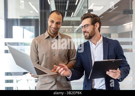 Two businessmen smile while working on a laptop in a modern office setting, discussing. One holds a clipboard. Collaboration is shown. Stock Photo