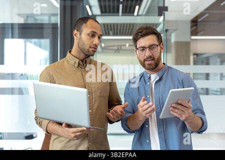 Two professional men collaborate in a modern office, using a laptop and tablet for discussion. They appear focused and engaged in their work. Stock Photo