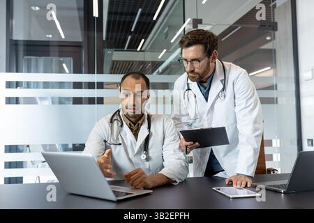 Two doctors in white coats are discussing something on a laptop, a clipboard, and a tablet in a modern office setting. Stock Photo