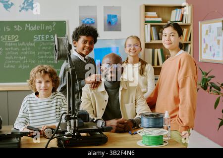 Multiracial schoolkids and teacher in modern classroom with 3D printer and spools of filament visible on table while standing beside blackboard featuring instructions Stock Photo