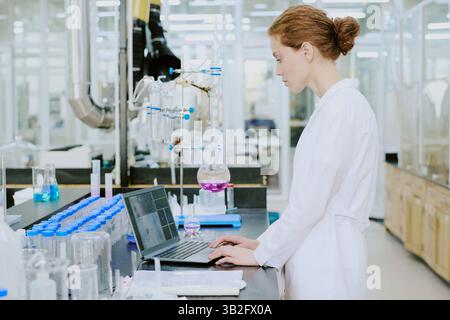 Scientist working in laboratory wearing white lab coat, using laptop for research. Laboratory setting with various scientific equipment and glassware visible in background Stock Photo
