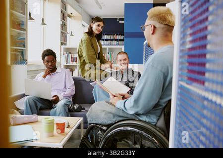 Diverse team members collaborating in modern office space, sitting comfortably, and using laptops and books. Inclusivity highlighted with wheelchair accessibility Stock Photo
