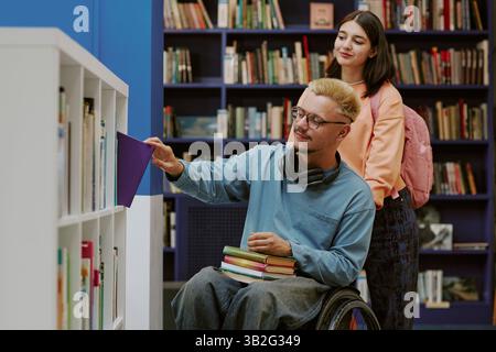 Two students exploring books in library, one in wheelchair perusing a new book while other standing and observing the process Stock Photo