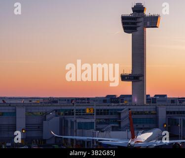 Air traffic control tower at JFK Airport, New York City, during a vibrant sunset with a commercial aircraft below at the gate. Stock Photo