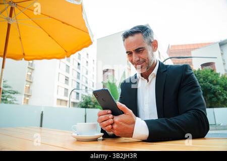 A focused businessman interacts with his smartphone, absorbed in its screen while sipping coffee at a cafe, showcasing the blend of technology and Stock Photo