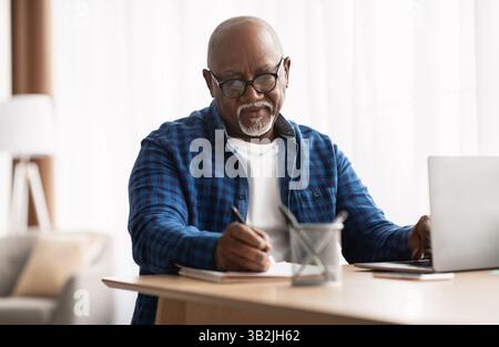 African American Man Taking Notes Sitting At Laptop In Office Stock Photo