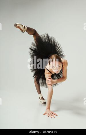 a ballerina in a black tutu is leaning forward leaning on her arm and lifting one leg up Stock Photo