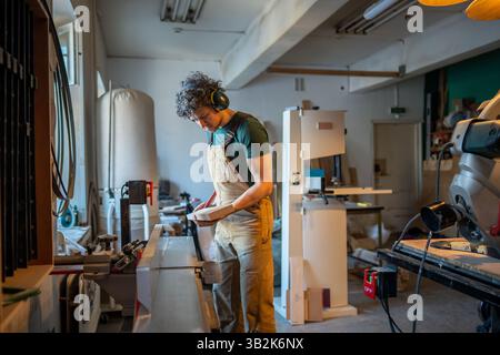Carpenter work on jointer machine in workshop, wear noise canceling earmuffs, handle wooden beam Stock Photo