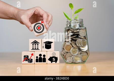 Glass jar filled with coins and a small seedling on a table. Highlights the concept of saving and financial growth. Wooden blocks with coins, Educatio Stock Photo