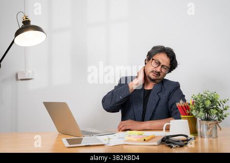 Health care concept A tired, uncomfortable Asian businessman sits at his desk in front of a computer, suffering from office syndrome, massaging his ne Stock Photo