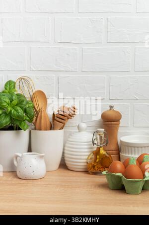 Kitchen counter displaying fresh eggs, basil plant, oil, spices, and various utensils against a white brick wall, embodying a home cooking atmosphere Stock Photo