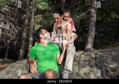 Parents and daughter taking a break during forest hike, sitting on a rock and sharing an affectionate moment. Symbol of connection, nature and balance Stock Photo