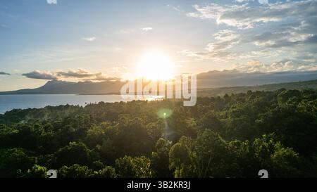 Aerial view of lush tropical forest meeting the ocean during sunset, with golden sunlight streaming through the clouds. Stock Photo