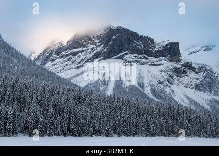 The tranquil winter landscape features towering snow-covered mountains reflecting sunlight while tall evergreen trees surround a frozen lake, creating Stock Photo