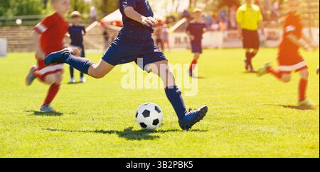 Children playing soccer in a match, with a player in blue kicking the ball while players in red chase, under sunny weather on a grassy field Stock Photo