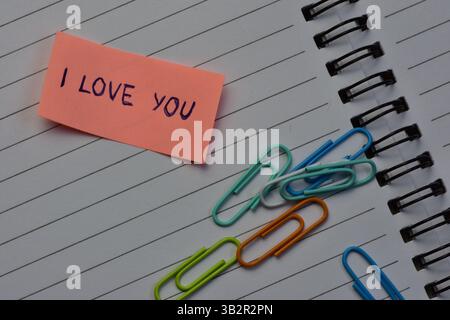 Message I love you on pink sticky note for valentines day next to various color paper clips on spiraled notebook open pages background Stock Photo