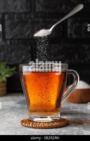 White sugar falling from teaspoon into glass cup of hot tea placed on coaster against black brick wall as background Stock Photo