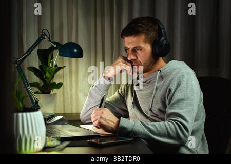 Focused man in headphones working on laptop at night and writing notes. Freelancer working remotely from home Stock Photo