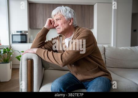 Lonely confused elderly man sits lost in thought staring blankly on home sofa, abandoned seniors Stock Photo