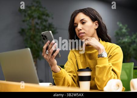 Young professional engages with her smartphone while surrounded by plants and a laptop. Stock Photo