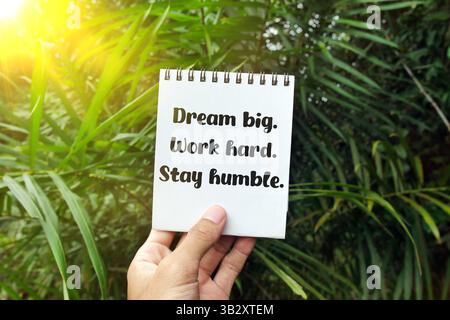 Hand holding notebook with text Dream big. Work hard. Stay humble in tropical green nature background with sunlight flare, encouraging motivation Stock Photo