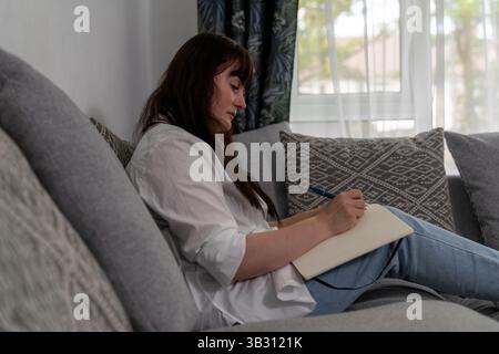Young woman jotting down notes in a notebook while relaxing on a comfortable sofa in a cozy living room filled with natural light Stock Photo