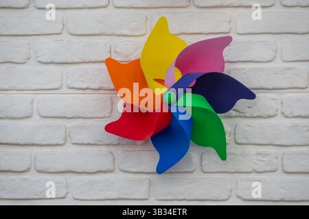 A colorful toy pinwheel with six vibrant blades—red, orange, yellow, pink, purple, blue, and green—is mounted against a white brick wall. Stock Photo
