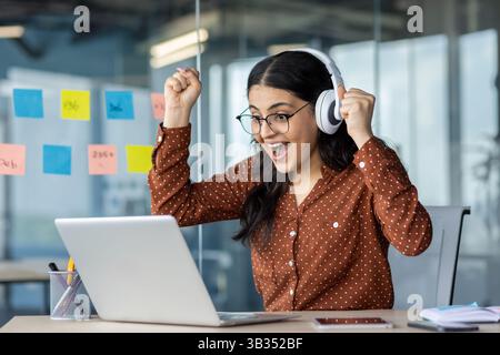 A joyful woman celebrates a win, wearing headphones and glasses, looking at her laptop in an office setting. She is excited. Stock Photo