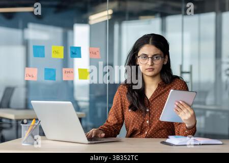 An Indian woman with glasses sits in an office, using a laptop and holding a tablet with sticky notes on the glass behind her. Stock Photo