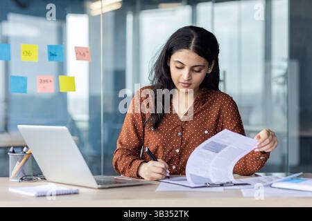 A focused woman, working intently on paperwork at a desk in a modern office setting. A laptop sits beside her, and sticky notes are visible. Stock Photo