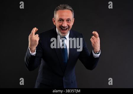 Joyful businessman in suit cheering with both hands raised and fingers crossed, expressing excitement and hope. Stock Photo