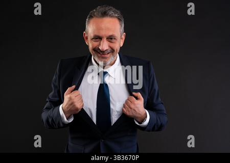 Mature businessman in suit smiling confidently and adjusting his jacket, in a playful and charismatic pose against a black background. Stock Photo