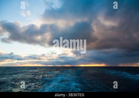 Sunset sky with rays of light from under the clouds in the ocean and traces on the water from the movement of the ship. Stock Photo