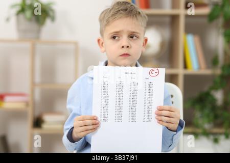 School grade. Sad boy holding answer sheet with letter C and minus symbol indoors Stock Photo