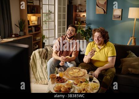Two friends sitting on a couch laughing while having pizza and snacks. Cozy living room setting with warm lighting and a friendly atmosphere Stock Photo