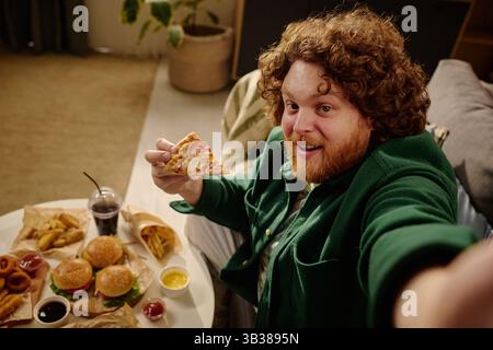 Young curly-haired man smiling widely while holding a slice of pizza, surrounded by various fast food items on a table. Cozy home setting with soft lighting Stock Photo