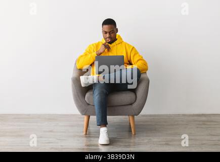 Black Man Using Laptop Sitting In Armchair Near Gray Wall Stock Photo
