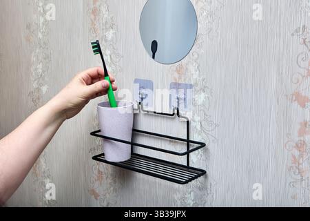 Hand places green toothbrush into light purple cup resting on black metal rack attached to wall with two transparent adhesive hooks. Stock Photo