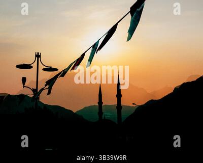 Dramatic sunset silhouettes a mosque with minarets against a backdrop of mountains and flags. Atmospheric sunset view over a mosque with two minarets. Stock Photo