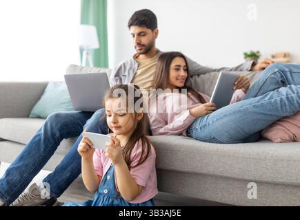 Gadgets addiction. Eastern family using different electronic devices, resting together in living room at home Stock Photo