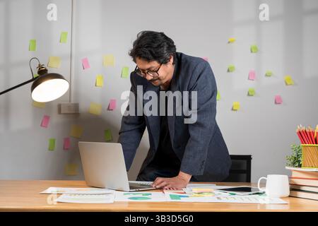 Serious millennial man using laptop sitting at the table in a home office, looking at the paper, communicating online, writing emails, distantly worki Stock Photo