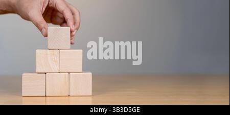 A hand places a wooden block atop a pyramid structure made of blocks, symbolizing strategy, planning, or construction in a professional setting, isola Stock Photo