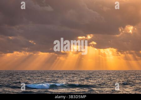 Sunrise over the ocean in Florida with golden sunrays breaking through dramatic clouds, illuminating the waves and peaceful sea horizon Stock Photo