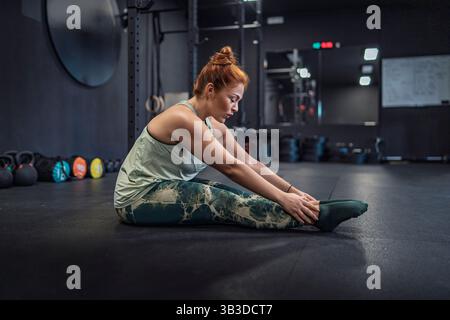 Young woman doing leg stretching exercises during warm-up in modern gym. Concept of fitness, health, and wellbeing. Stock Photo