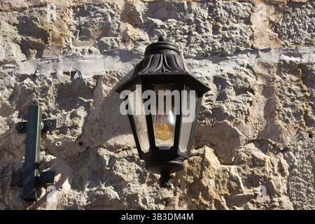 Antique-style black metal street lamp against natural stone wall. Vintage wrought iron lamp post against stone brick wall background. Stock Photo