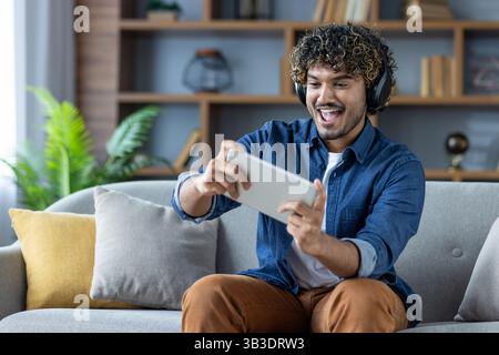 A young man with curly hair sits on a couch, engrossed in playing a game on his tablet. He wears headphones and appears excited. Stock Photo